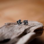 A pair of sterling silver stud earrings shaped like tiny owls, perched on driftwood with a warm golden background.